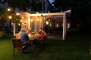 Family sitting at a table at night in the backyard under a white pergola with hanging lights